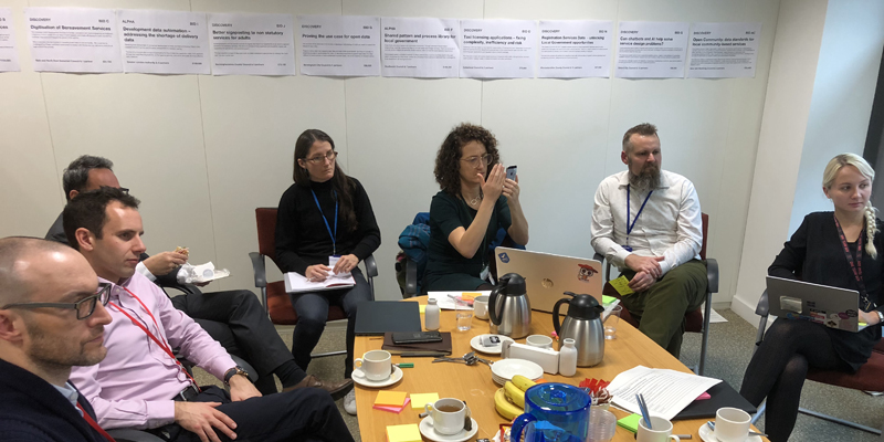 7 members of staff sit around a table, listening to another staff member out of picture. Print outs of each full application for the Local Digital Fund are printed and stuck to the wall behind them.