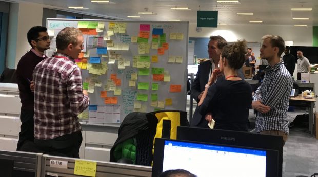 Sheldon and 4 other team members stood around a whiteboard covered in post-it notes. The team are taking part in a regularly held stand-up meeting.