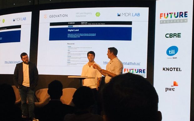 3 male speakers on stage at the event, with the Digital Land website being shown on the screen behind them. The image was provided by @Jessinblue on Twitter.