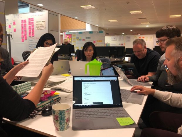 7 members of the Local Digital Collaboration Unit sat around a table, assessing Local Digital Fund applications on paper and laptops.