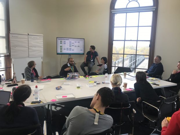 11 members of the Local Digital Collaboration Unit sit around a large table, and are looking at a presentation on a screen.