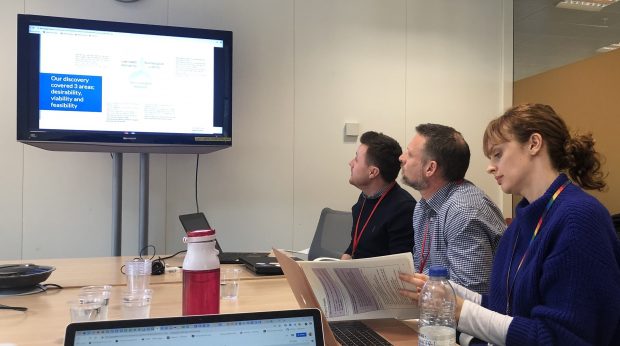 A photo from an interview in round 4 of the Local Digital Fund. 3 people sit on the right hand side of a desk, looking at a presentation on a tv screen at the end of the desk.