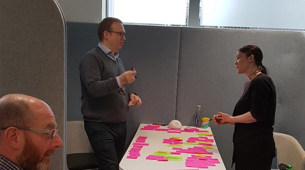 3 members of the pre-discovery team discuss at the kick off workshop, with a table of post-it notes between them.