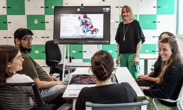 A group of colleagues having a meeting. One of them in standing next to a screen that shows some data charts.