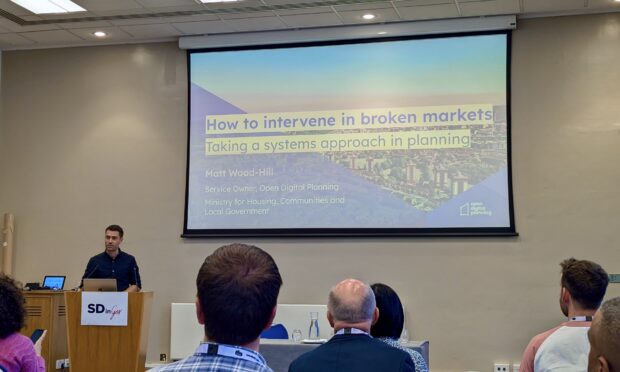 Matt Wood-Hill on stage - the slide being presented reads 'How to intervene in broken markets: taking a systems approach in planning' 