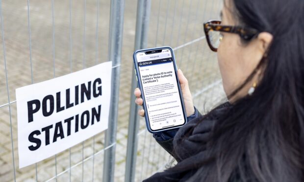 a person standing outside a gate with a 'polling station' sign and holding a phone showing a GOV.UK page which is not legible