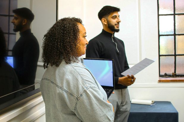 Two people standing up and smiling: one holding a laptop, the other one holding a piece of paper. 